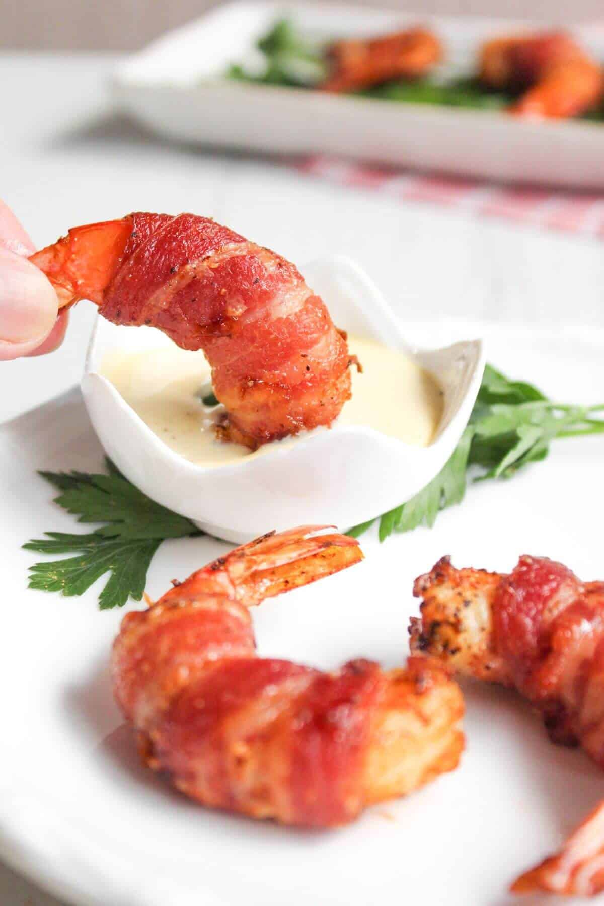 Bacon wrapped shrimp on a white plate with one dipped in sauce.
