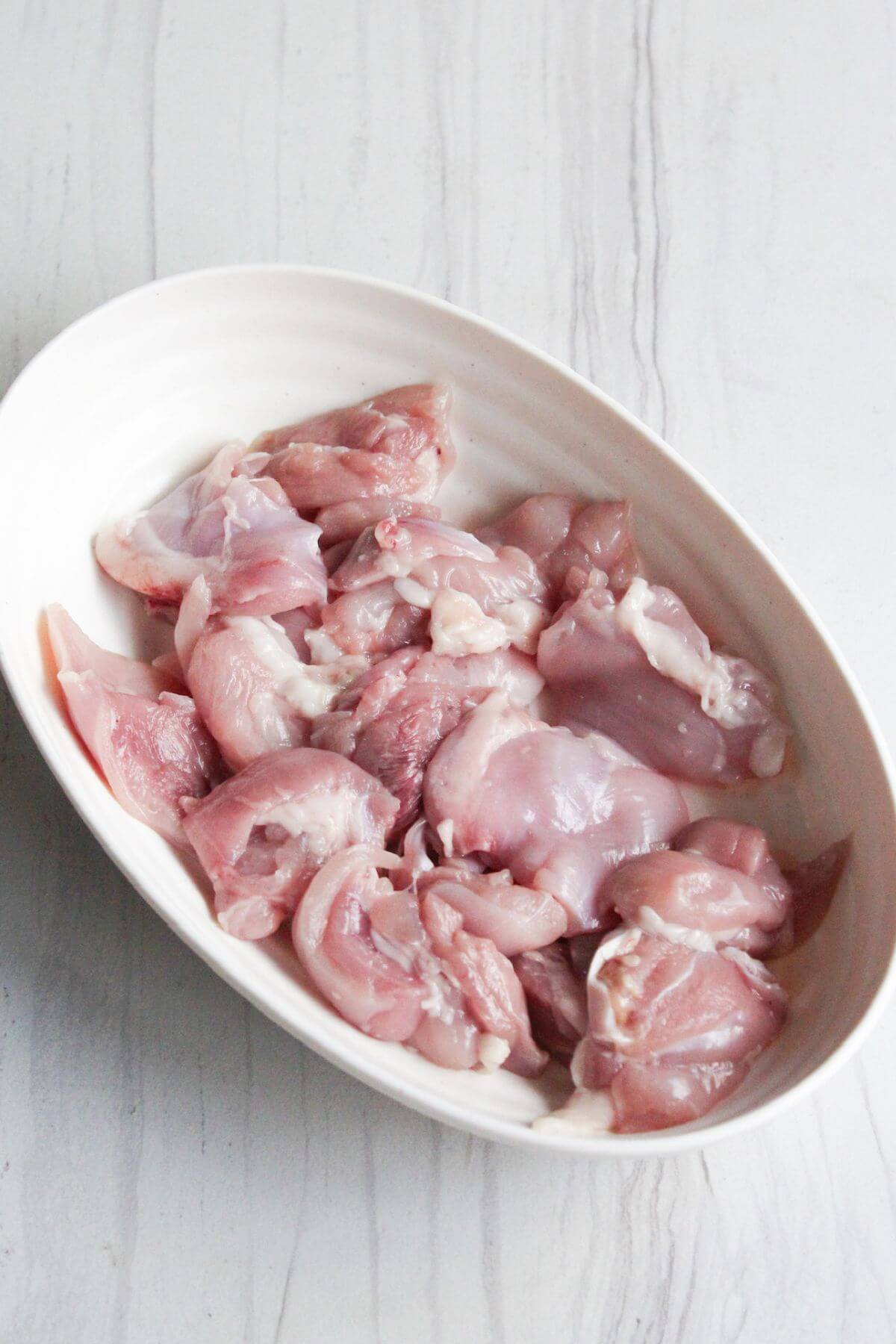 Cut raw chicken thighs in a white bowl on a table.