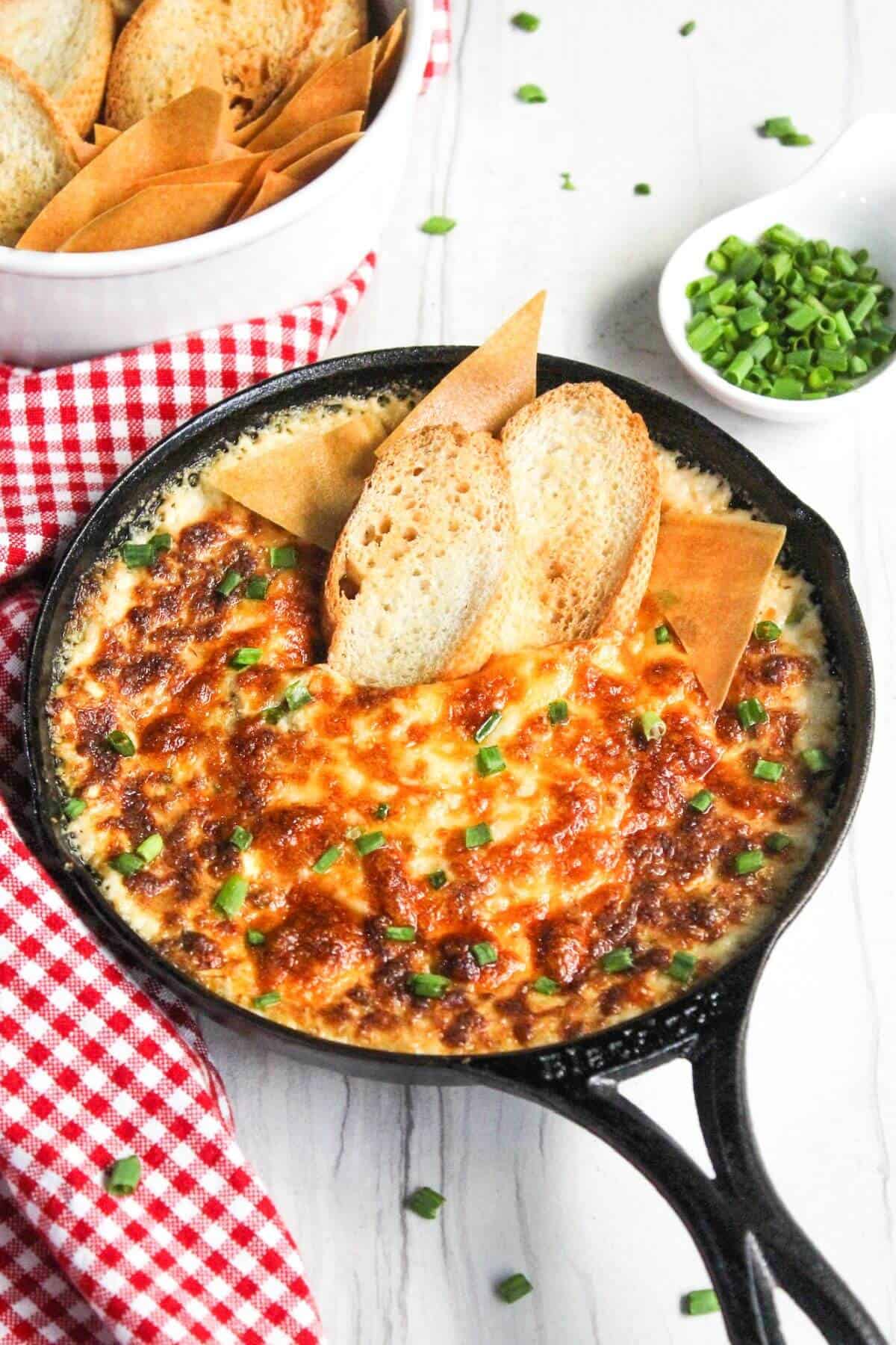 A skillet with cheesy crab dip and bread on a red and white checkered tablecloth.