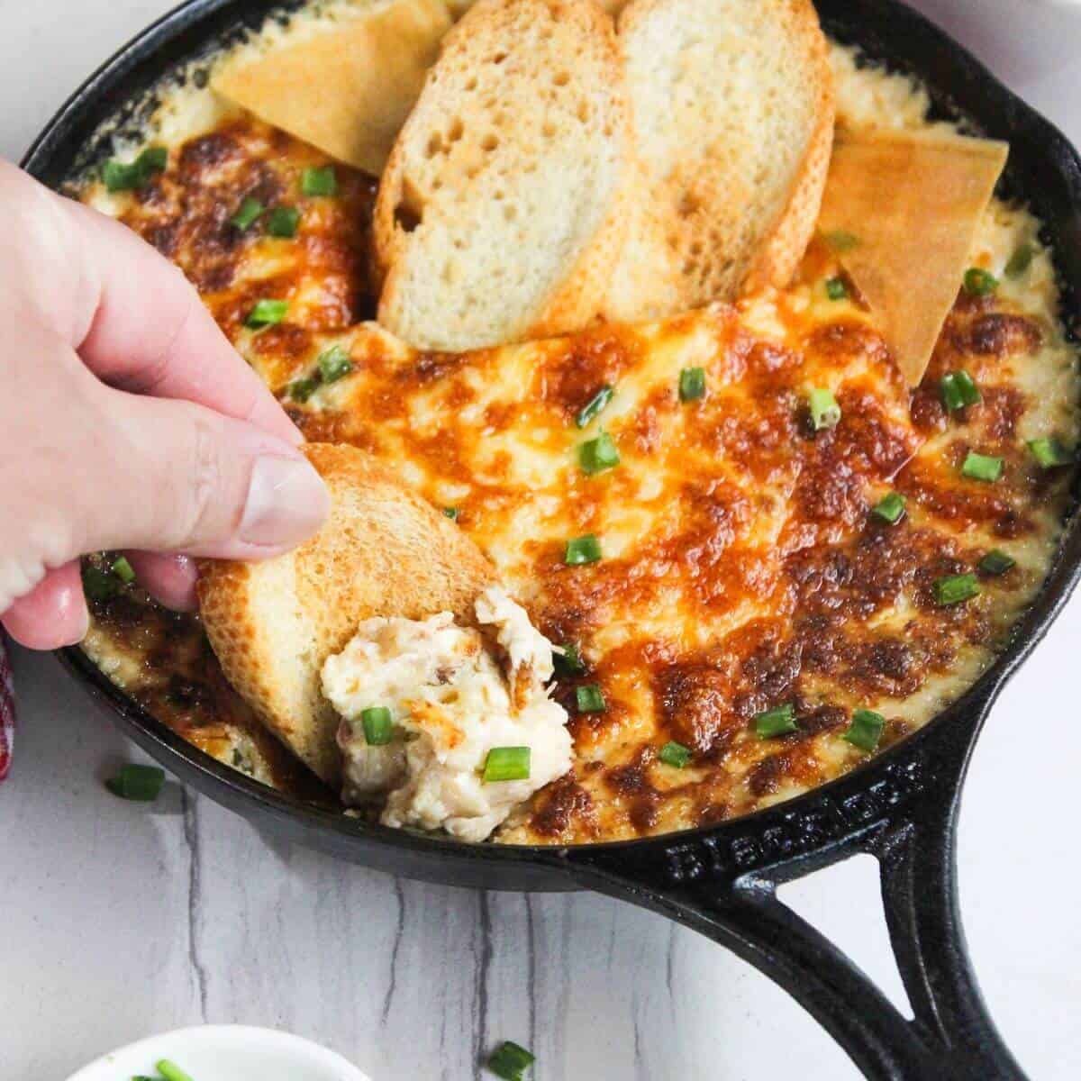 A person dipping bread into a skillet of cheesy crab rangoon dip.
