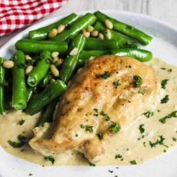 Cream cheese chicken breast in creamy sauce garnished with parsley, served with green beans topped with pine nuts on a white plate.