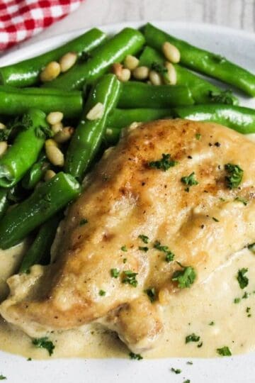 Cream cheese chicken breast in creamy sauce garnished with parsley, served with green beans topped with pine nuts on a white plate.