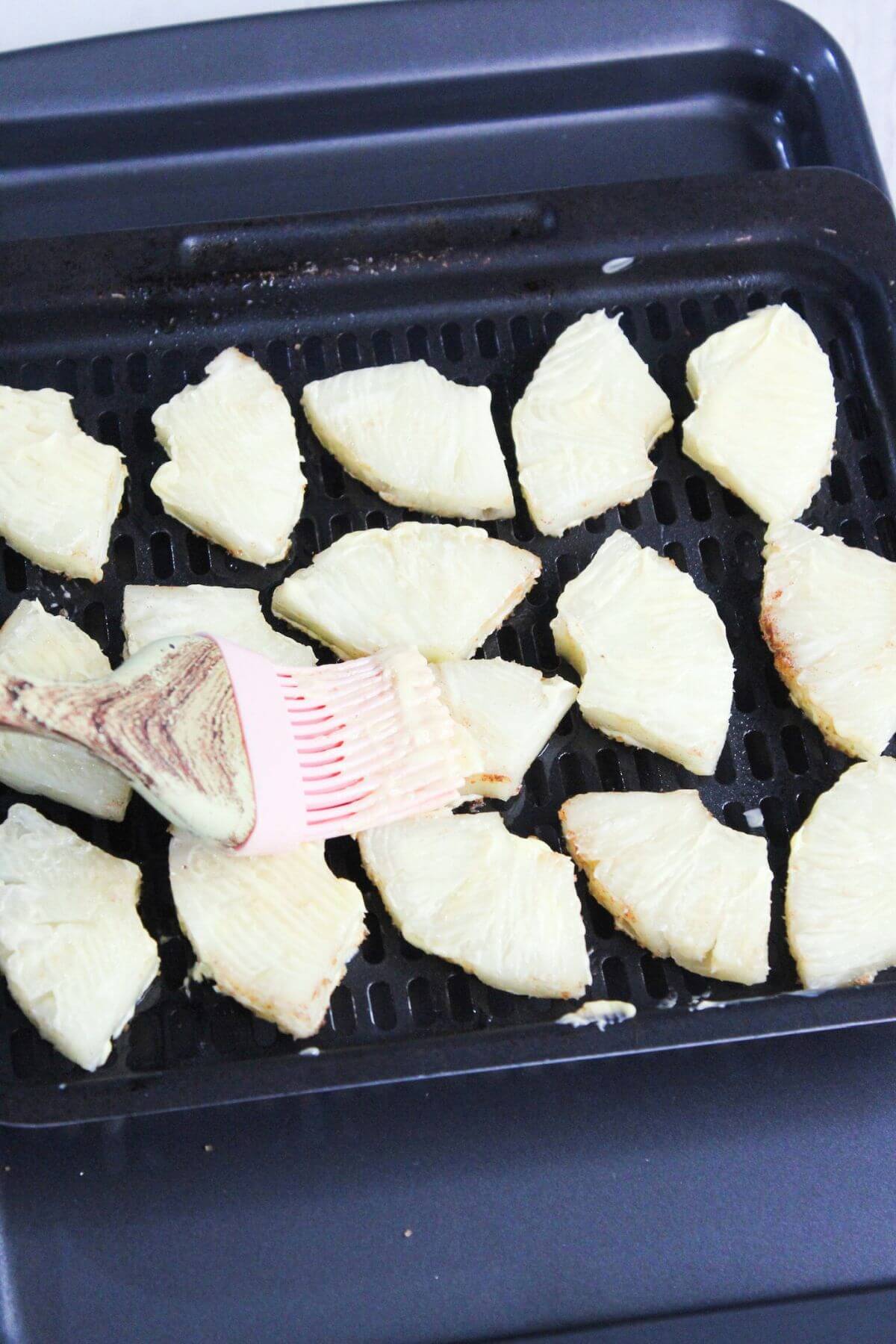 Sliced pineapple on an air fryer tray being brushed with butter.