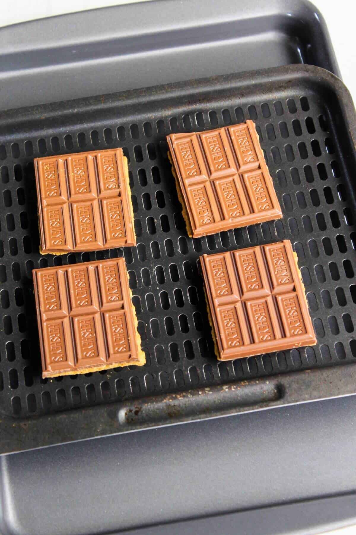 Four chocolate bars over graham crackers on air fryer tray.