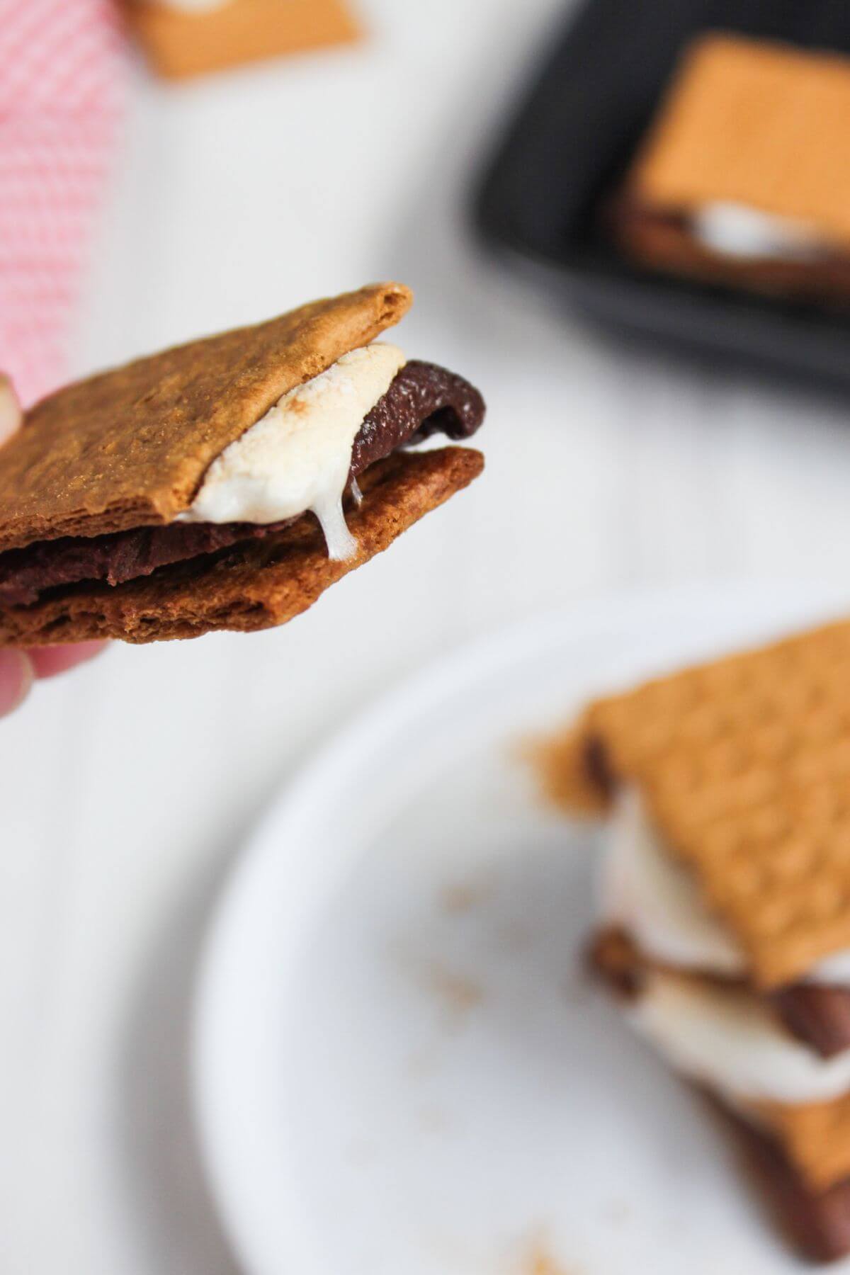 A person holding a s'more over a plate of s'mores.