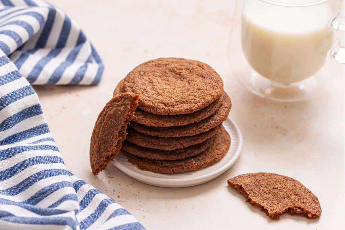 A stack of crisp brown sugar cookies on a plate with one cookie broken in half, accompanied by a glass of milk and a striped napkin.