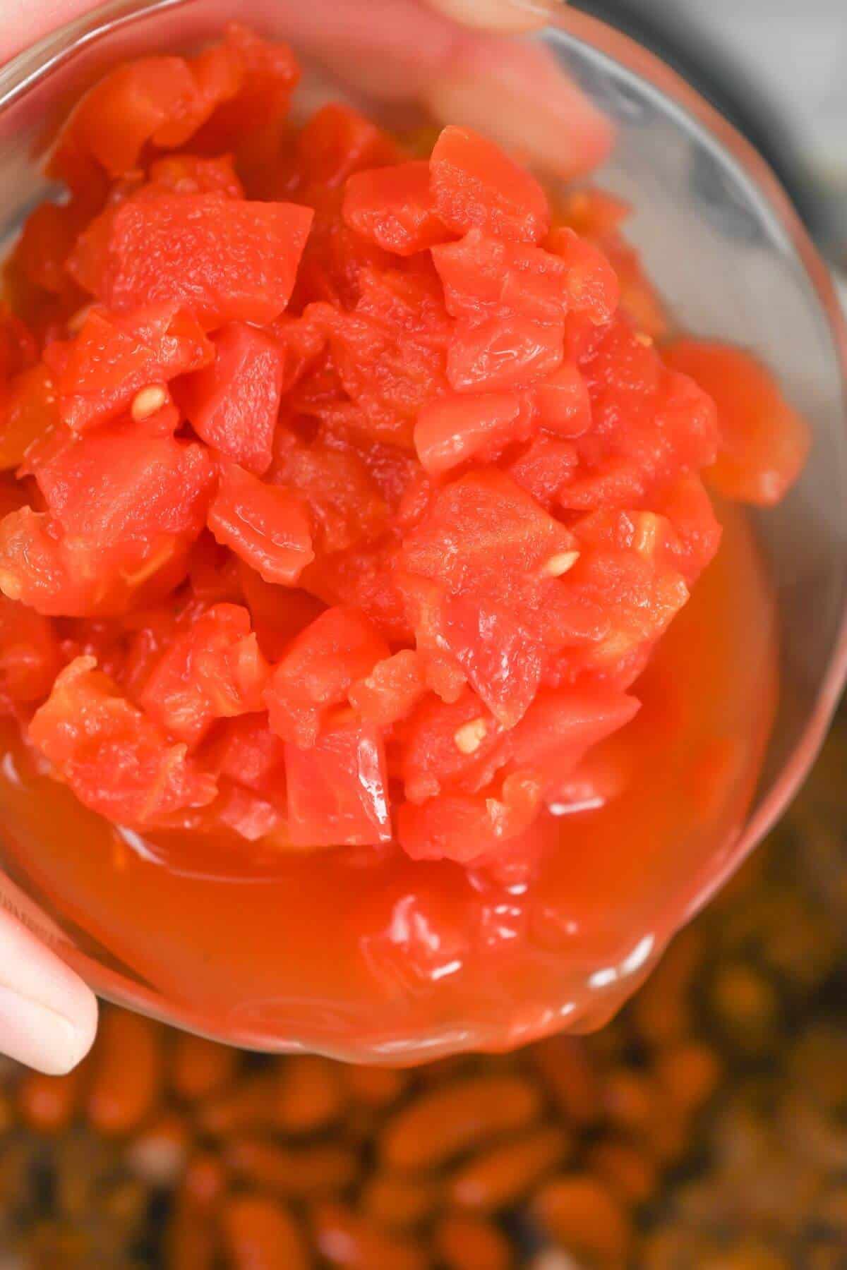 A hand holding a bowl of diced tomatoes over skillet of chili mix.
