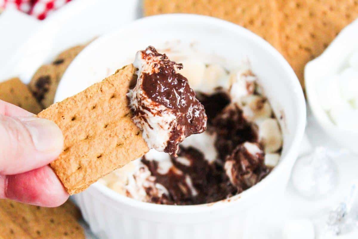 A person holding a graham cracker over a bowl of chocolate and marshmallows.