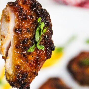 A piece of air fried chicken thigh on a fork, with a caramelized exterior and garnished with a small leaf, in focus against a blurred background of more chicken pieces.
