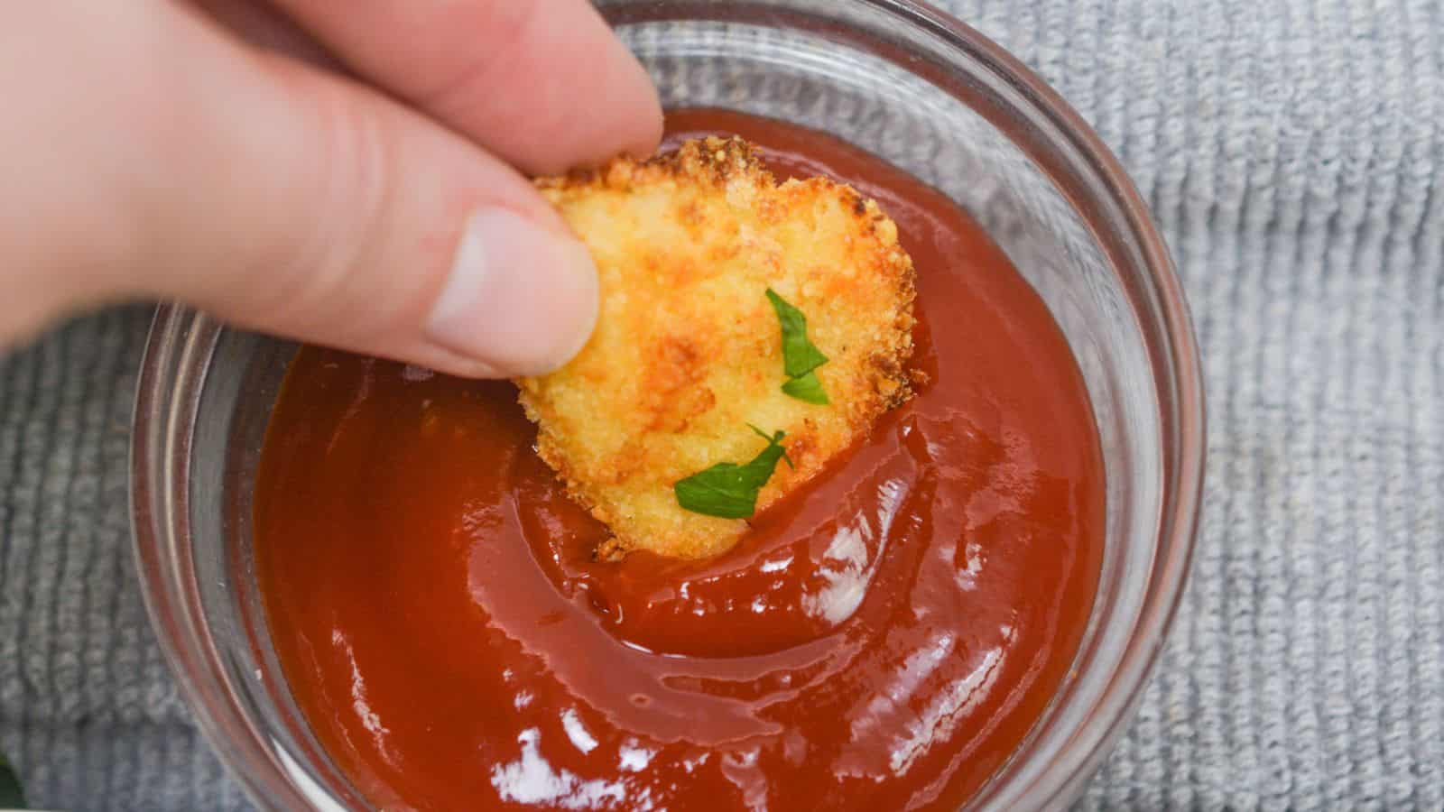 Hand dipping a chicken nugget into a bowl of ketchup, with a gray cloth background.