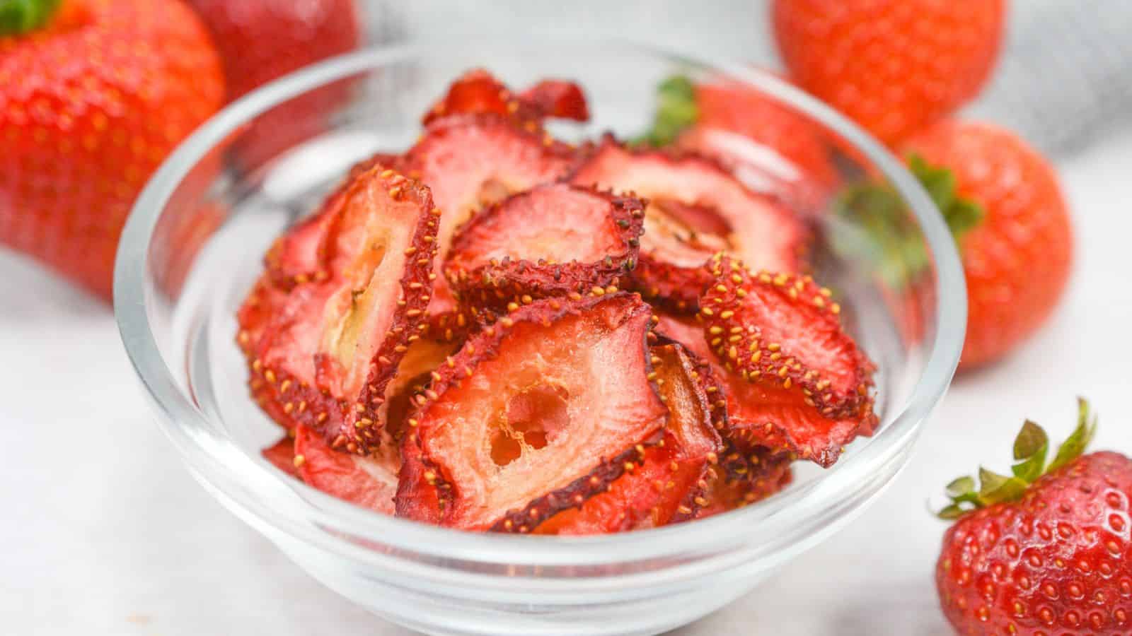 A bowl of sliced dried strawberries on a table, with fresh whole strawberries scattered around it.