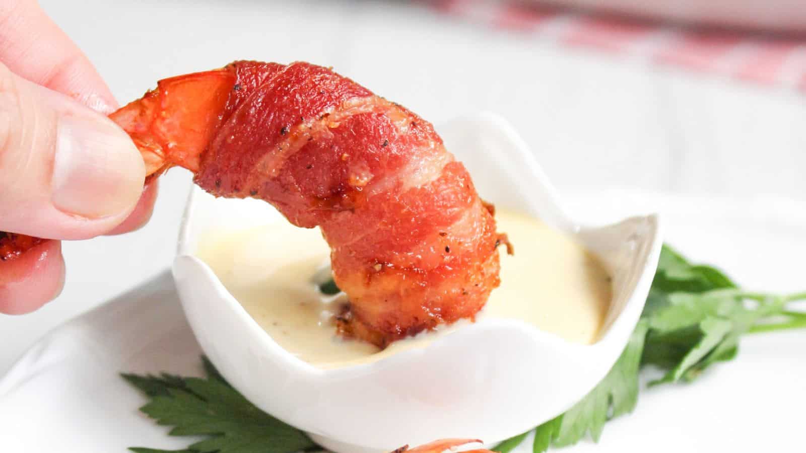 A hand holding a bacon-wrapped shrimp dipped into a small bowl of sauce, placed on a white surface with green herbs.