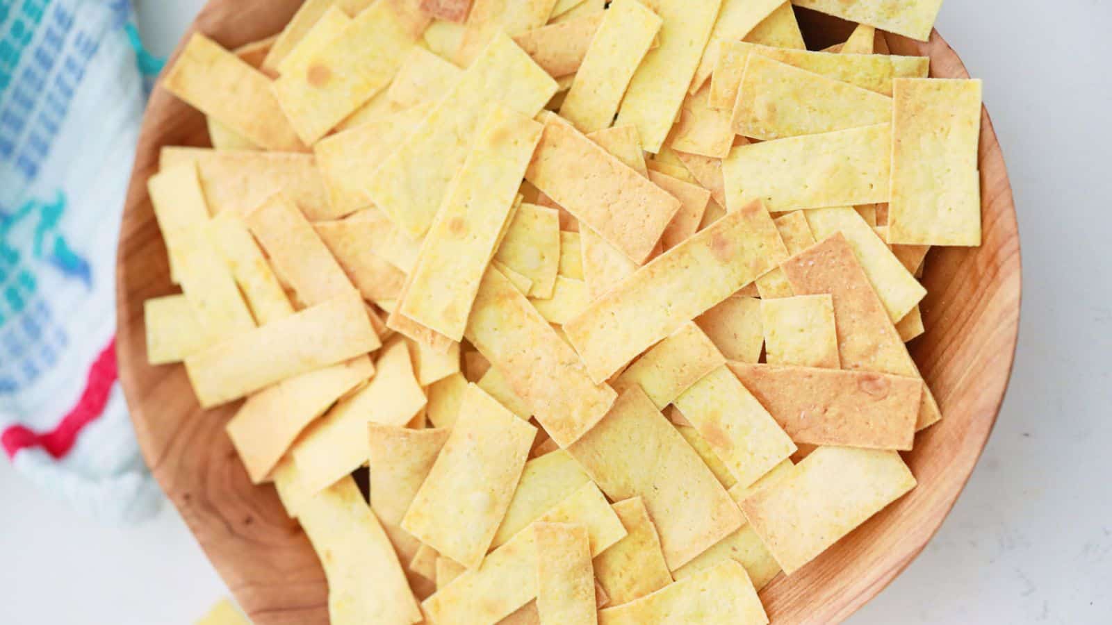 A wooden bowl filled with homemade rectangular yellow corn tortilla chips on a white surface.