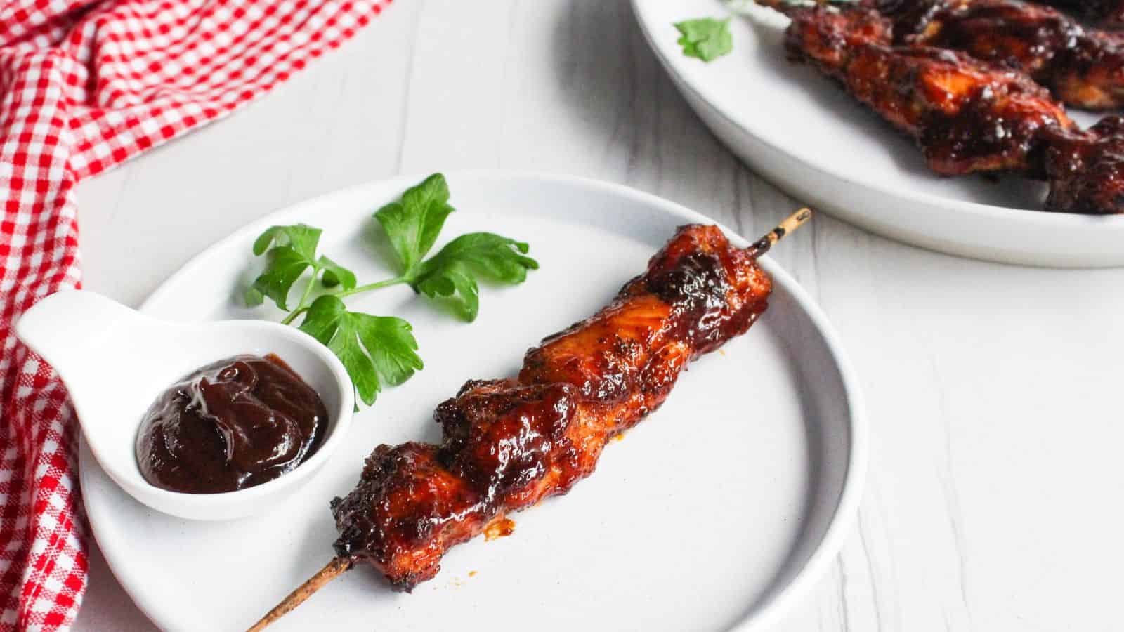 A grilled chicken skewer on a white plate with barbecue sauce and fresh parsley, beside a plate of more chicken skewers.