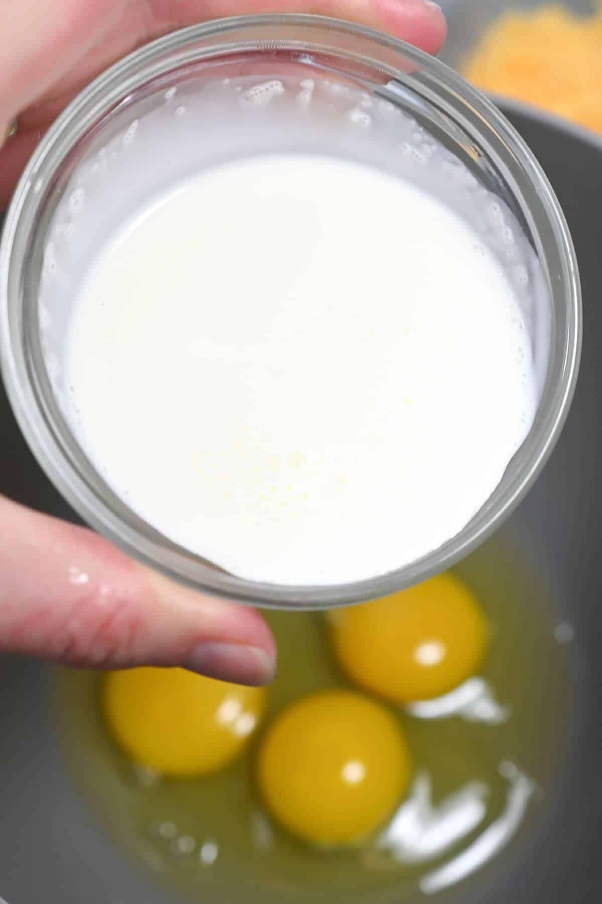 A person holding a glass of cream above a bowl containing eggs.
