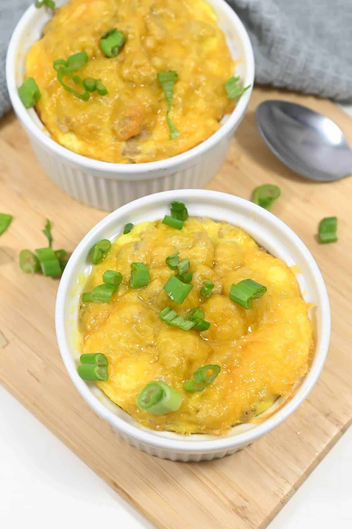 Two ramekins of baked cheesy biscuit breakfast casserole topped with green onions on a wooden surface.