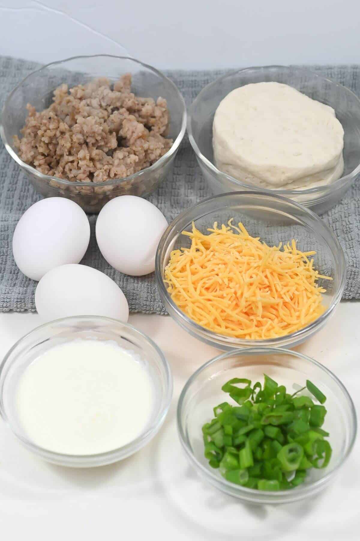 Ingredients for breakfast casserole laid out on a table, including ground meat, eggs, shredded cheese, chopped green onions, and cream.