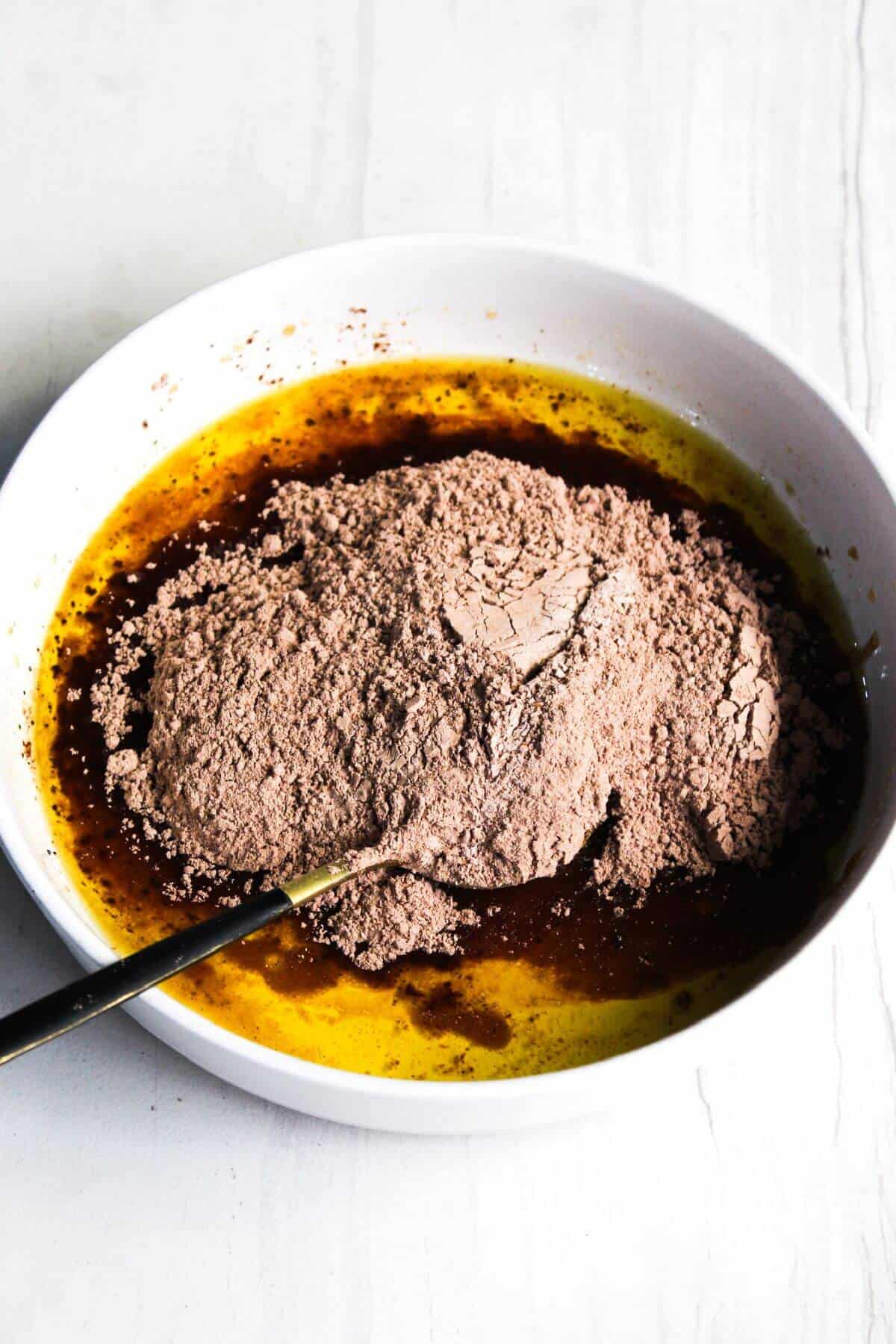 A bowl containing a mixture of melted butter and cocoa powder with a metal spoon on a white background.