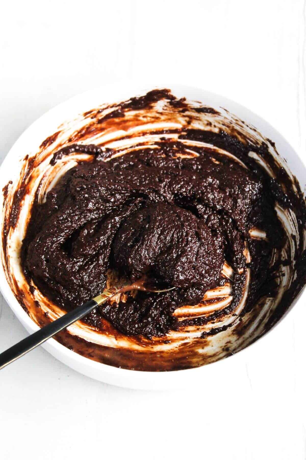 A bowl of thick, dark chocolate batter with a spoon, partially mixed with visible streaks of unmixed batter.