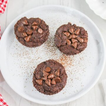 Three brownie bites topped with chocolate chips on a white plate, sprinkled with cocoa powder, beside a red checkered napkin.
