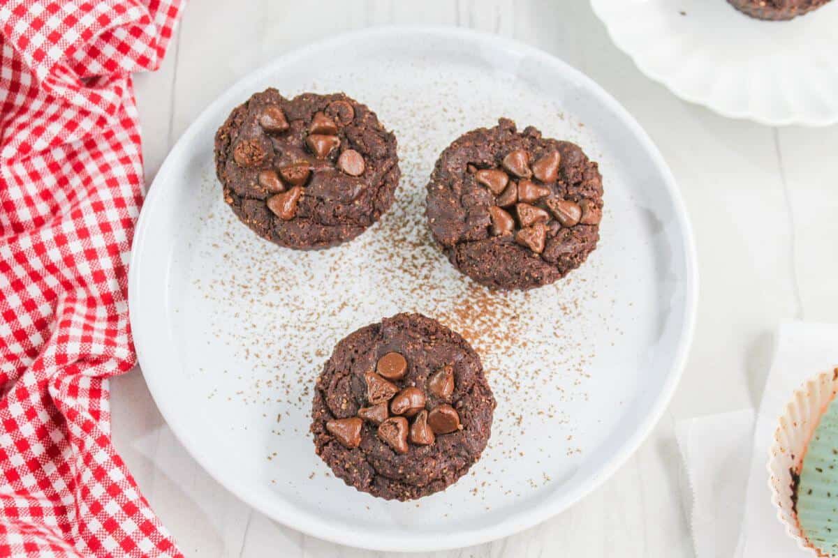 Three brownie bites topped with chocolate chips on a white plate, sprinkled with cocoa powder, beside a red checkered napkin.
