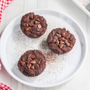 Three chocolate brownie bites topped with chocolate chips on a white plate, sprinkled with cocoa powder, with a red and white napkin on the side.