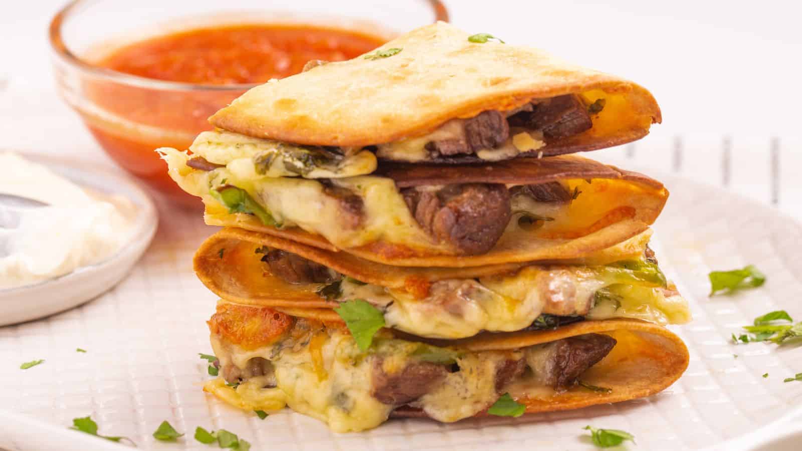 Stack of quesadillas filled with cheese and meat, garnished with herbs, served with dips on a white plate.