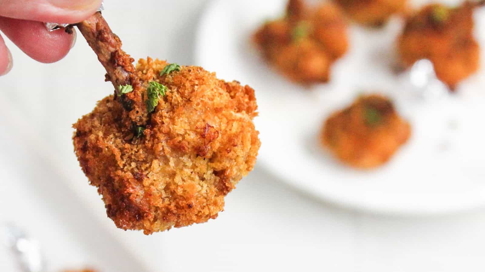 A close-up of a hand holding a crispy, breaded chicken drumette with more drumettes in the background on a white plate.
