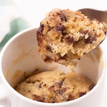 A spoon lifting a bite from a chocolate chip mug cake inside a white mug.