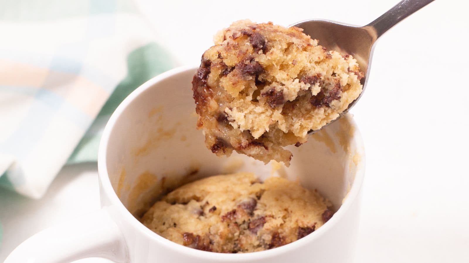 A spoon lifting a bite from a chocolate chip mug cake inside a white mug.