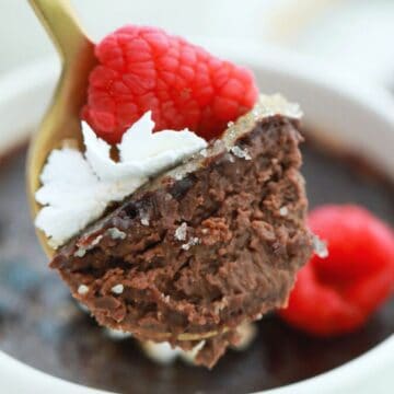 A spoonful of chocolate cream brulee garnished with a raspberry and whipped cream, above a white bowl containing more dessert.