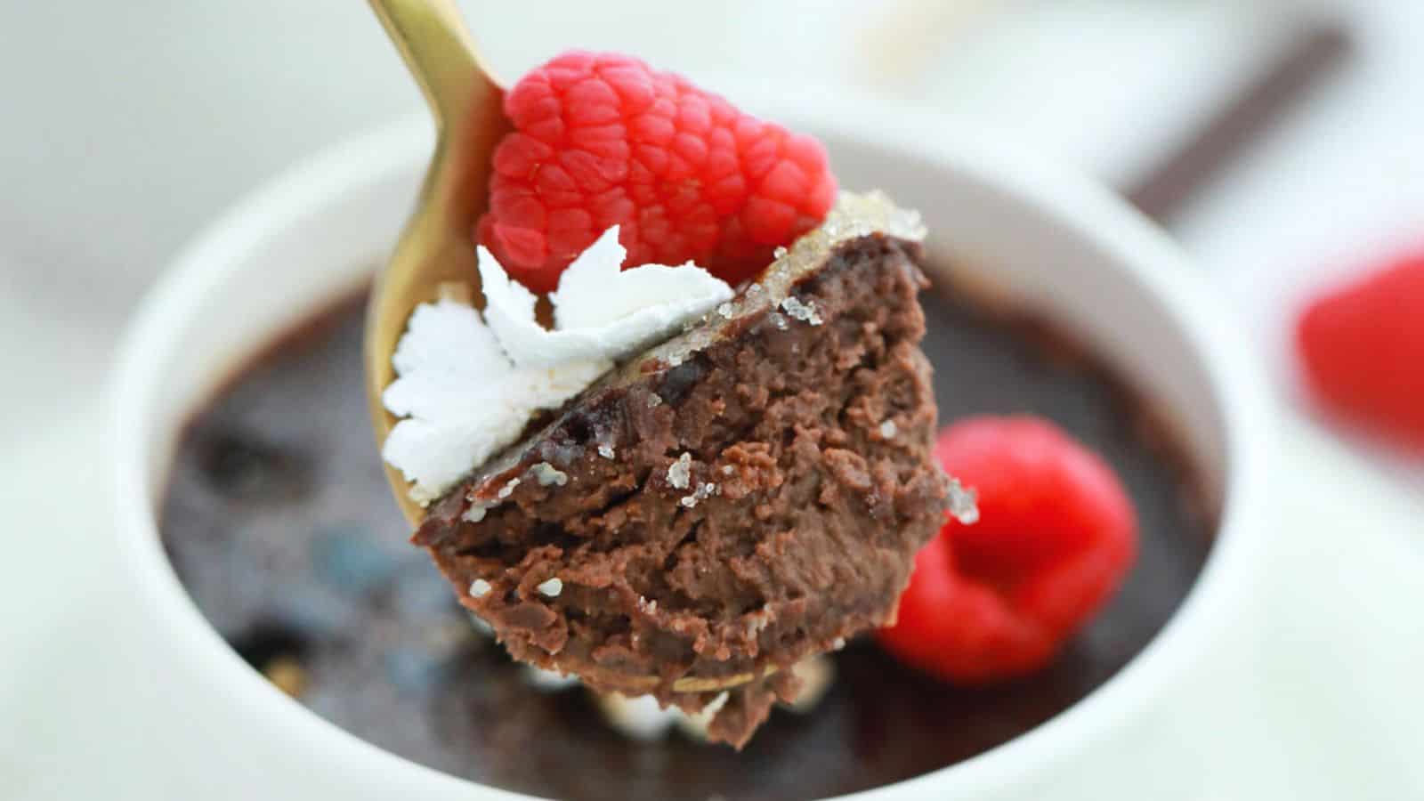 A spoonful of chocolate cream brulee garnished with a raspberry and whipped cream, above a white bowl containing more dessert.