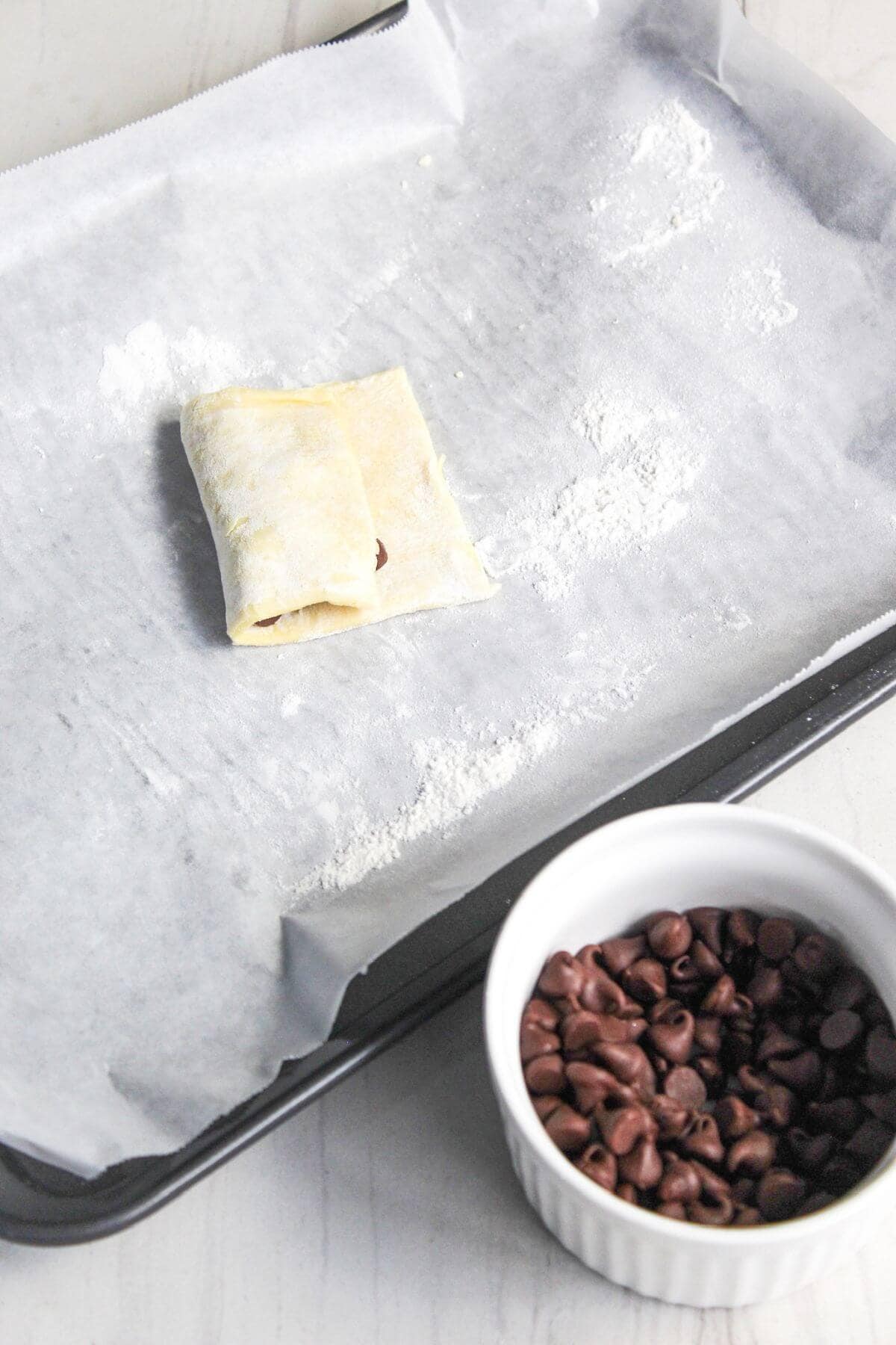 Uncooked filled puff pastry on a baking sheet with parchment paper, next to a small bowl of chocolate chips.