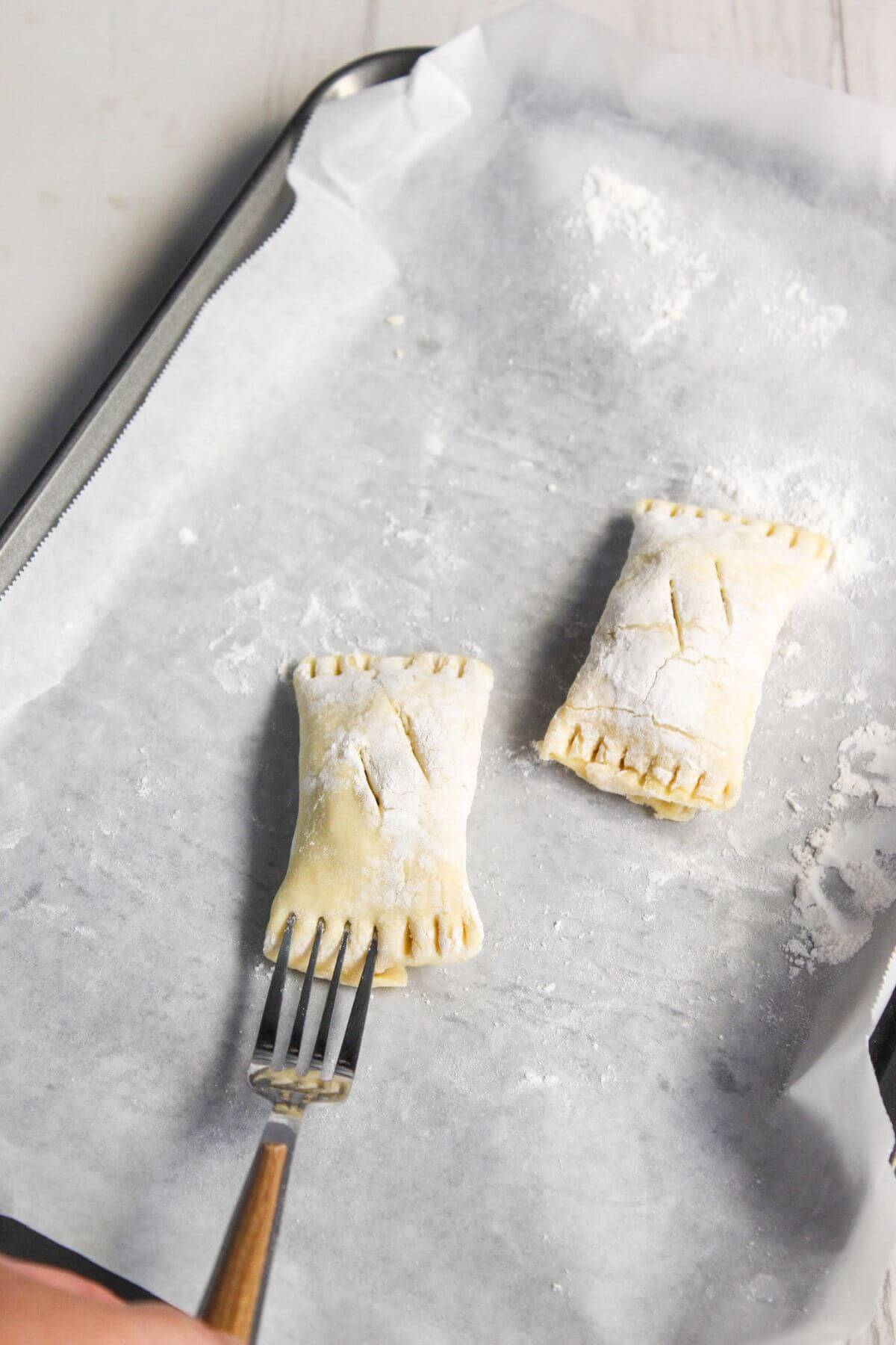 Two uncooked pastries on parchment-lined baking tray, with a hand holding a fork pressing edges of one pastry.