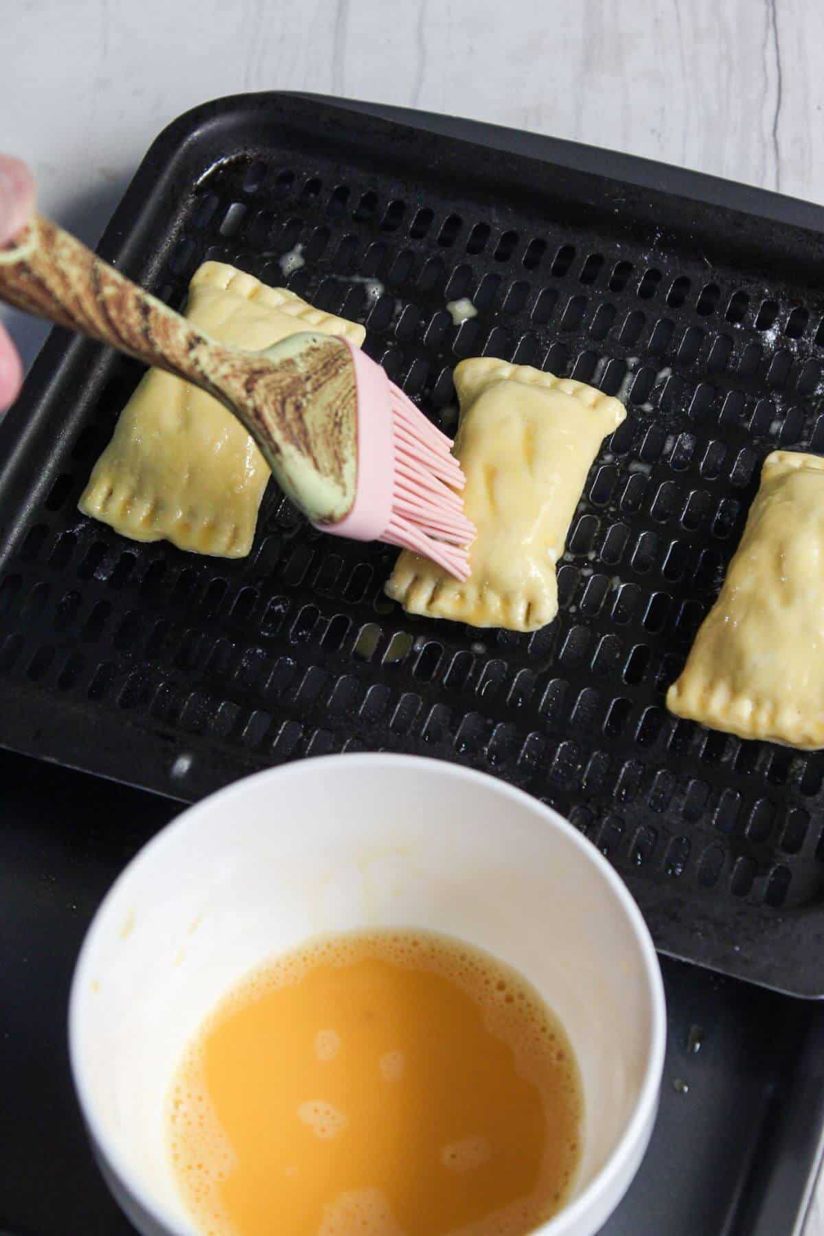 A person brushes egg wash on pastries on a baking tray, with a bowl of egg wash nearby.