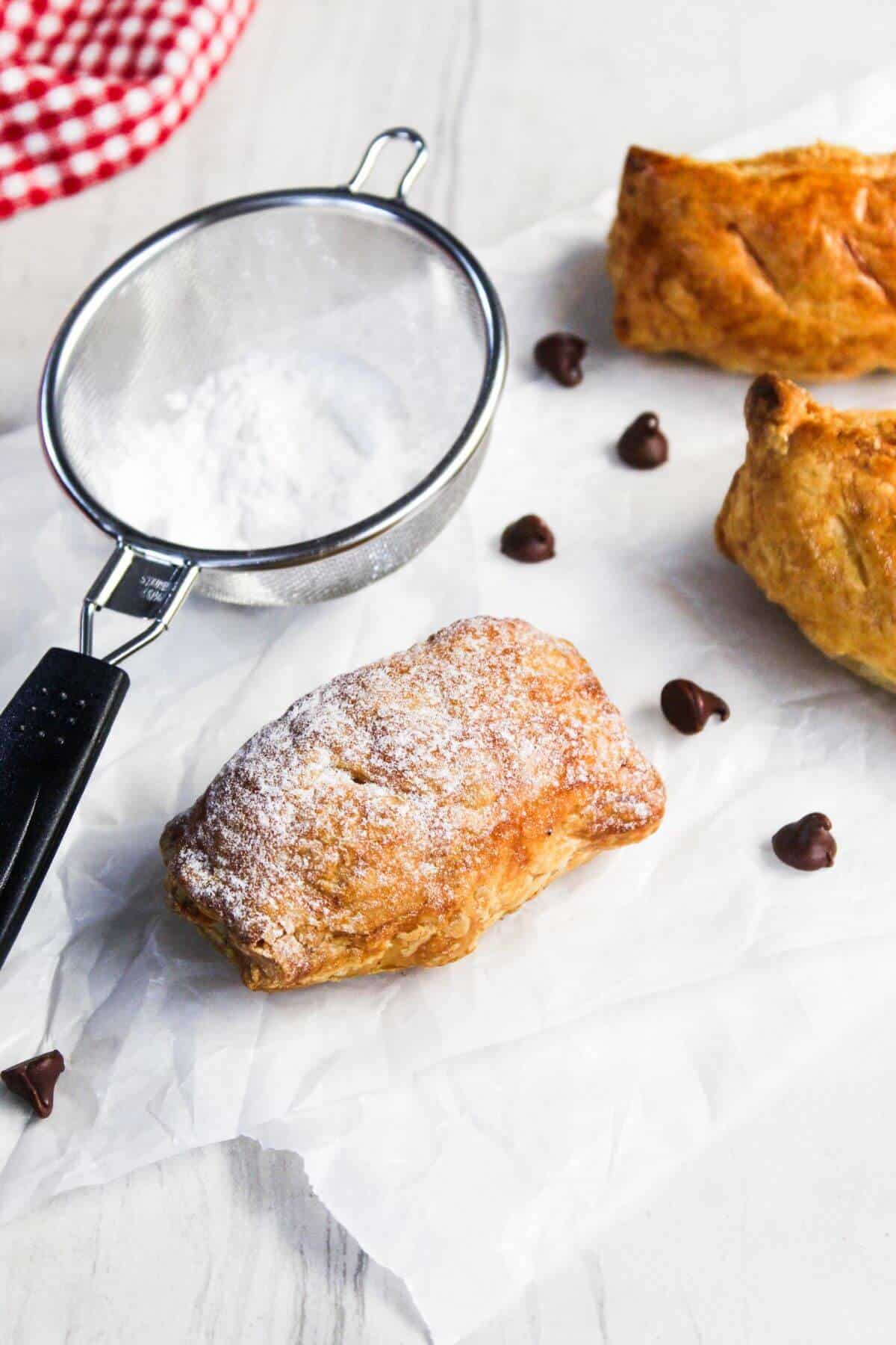 Freshly baked chocolate-filled pastry dusted with powdered sugar on a white surface, with a sieve and extra chocolate chips nearby.