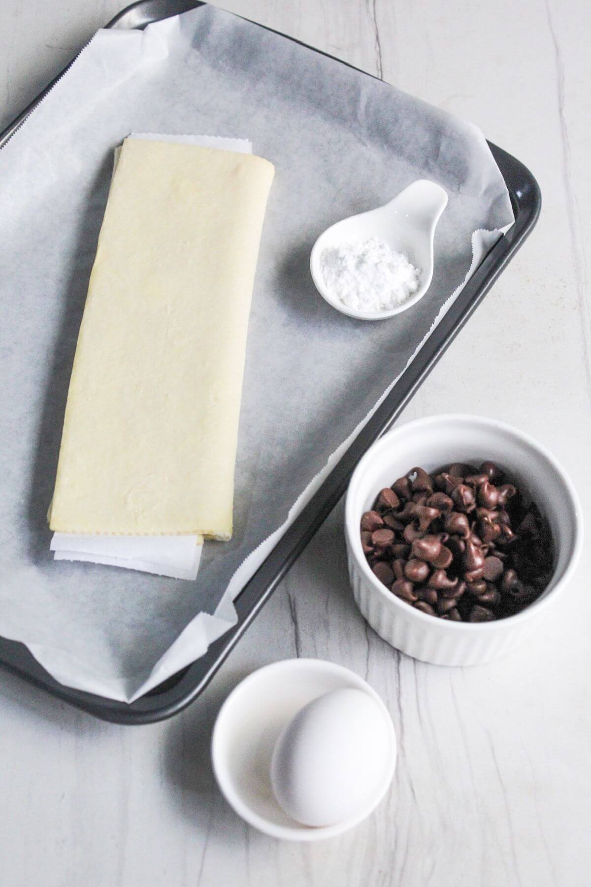 A baking tray with a sheet of puff pastry, a bowl of chocolate chips, an egg, and a spoonful of flour on parchment paper.