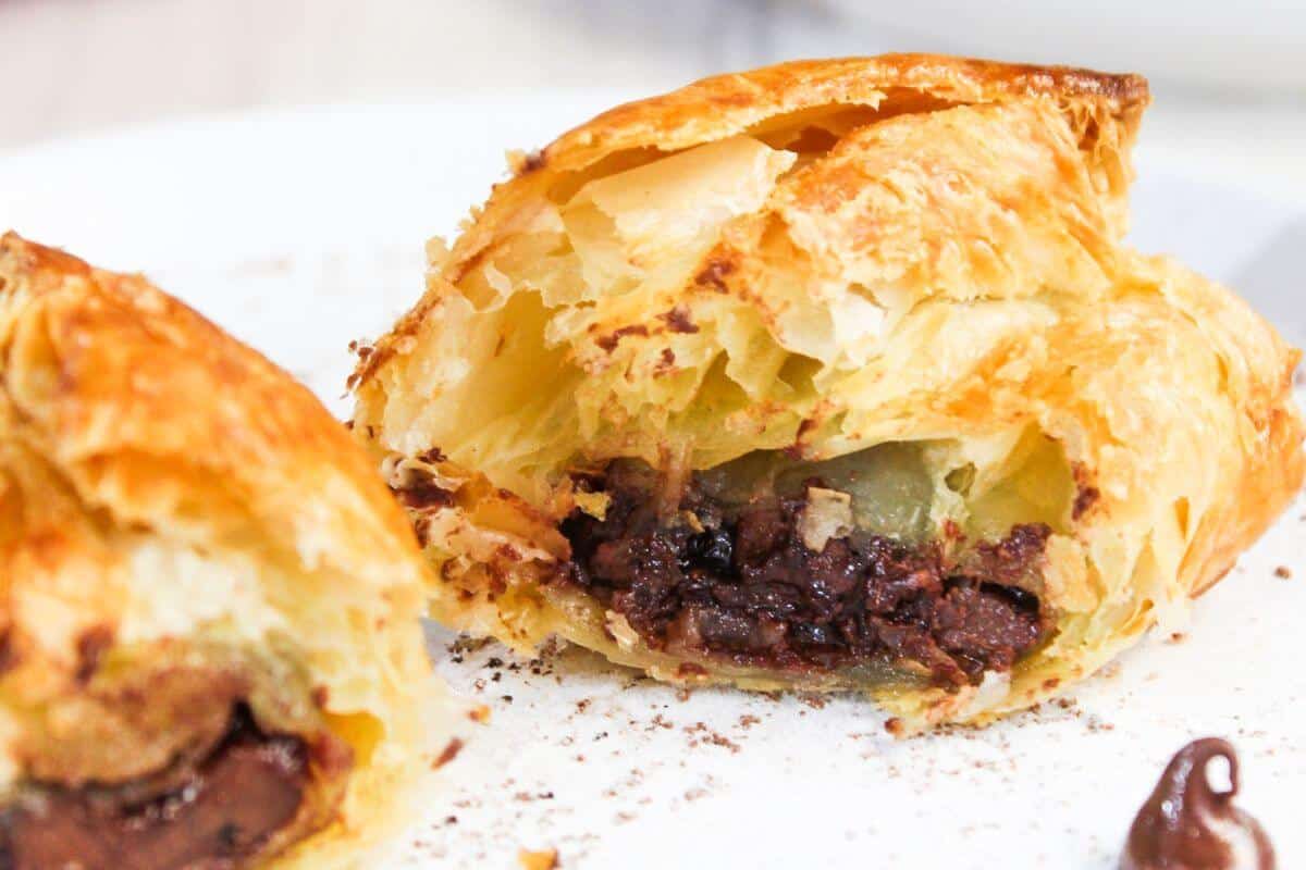 A close-up of a pastry cut open to reveal a filling of chocolate, served on a white plate with a smear of chocolate sauce.