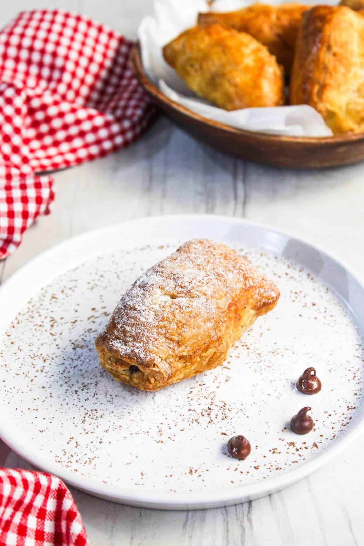 A freshly baked chocolate pastry dusted with powdered sugar on a white plate, decorated with chocolate chips and served on a wooden table with a red checkered napkin.