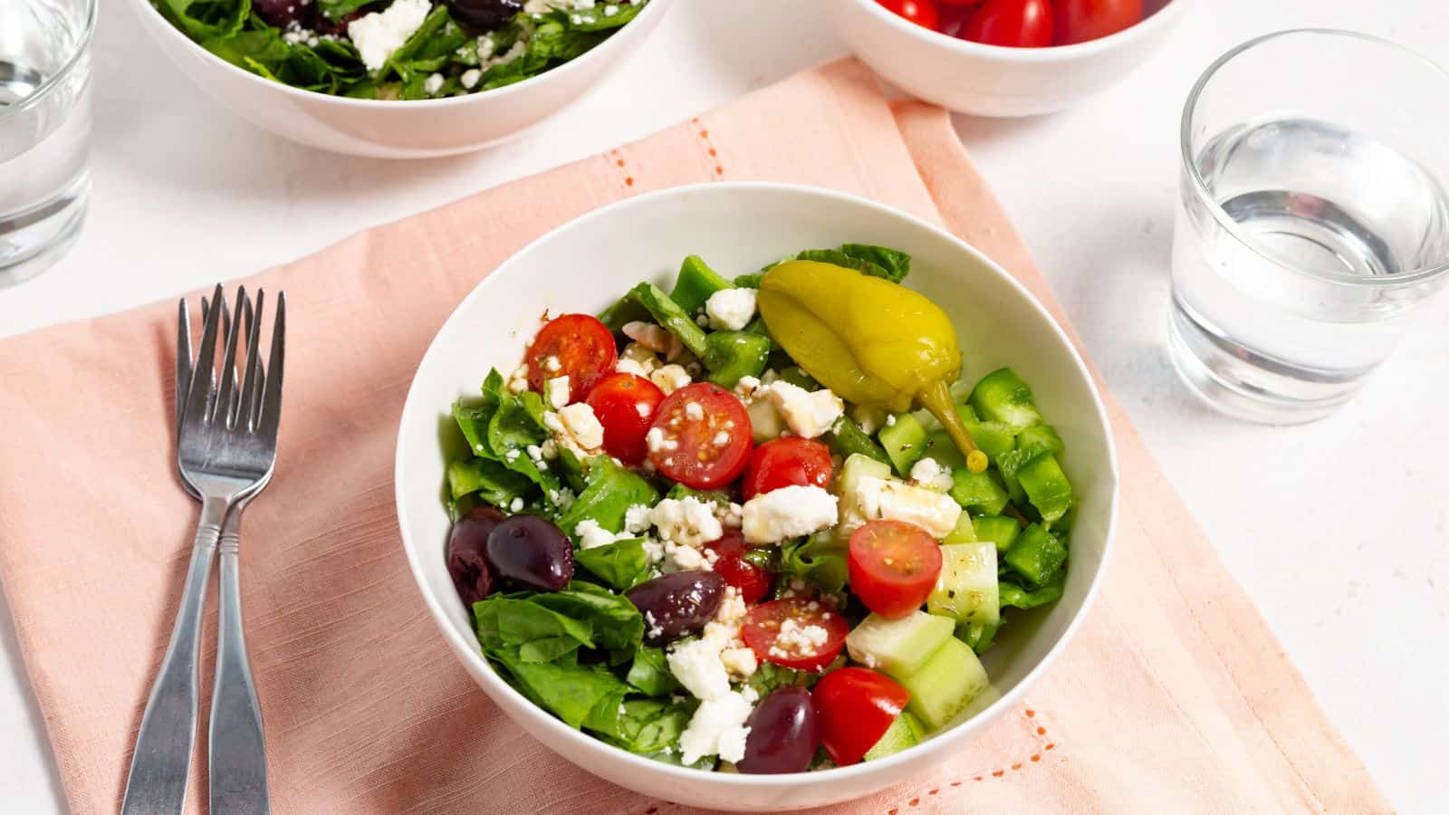 A fresh greek salad with tomatoes, feta cheese, cucumbers, olives, and peppers, served with a fork and a glass of water on the side.