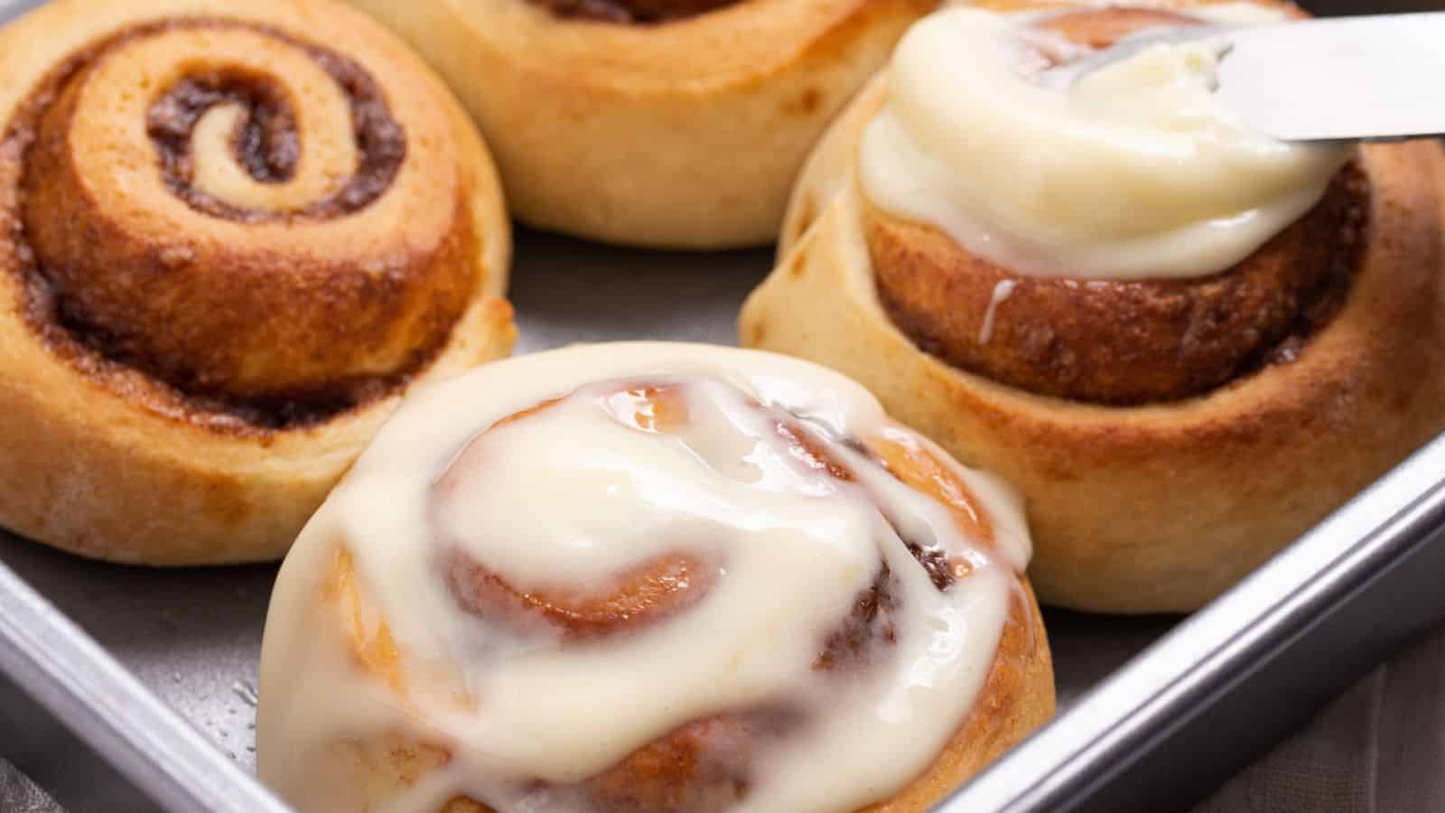 A person spreads white frosting on a freshly baked cinnamon roll in a tray with others nearby.