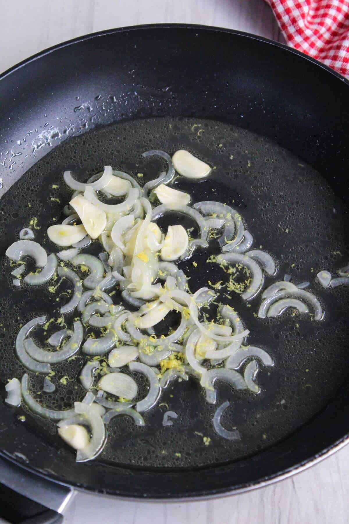 Sliced garlic and onions sautéing in oil in a black frying pan on a stove, with visible sizzling.