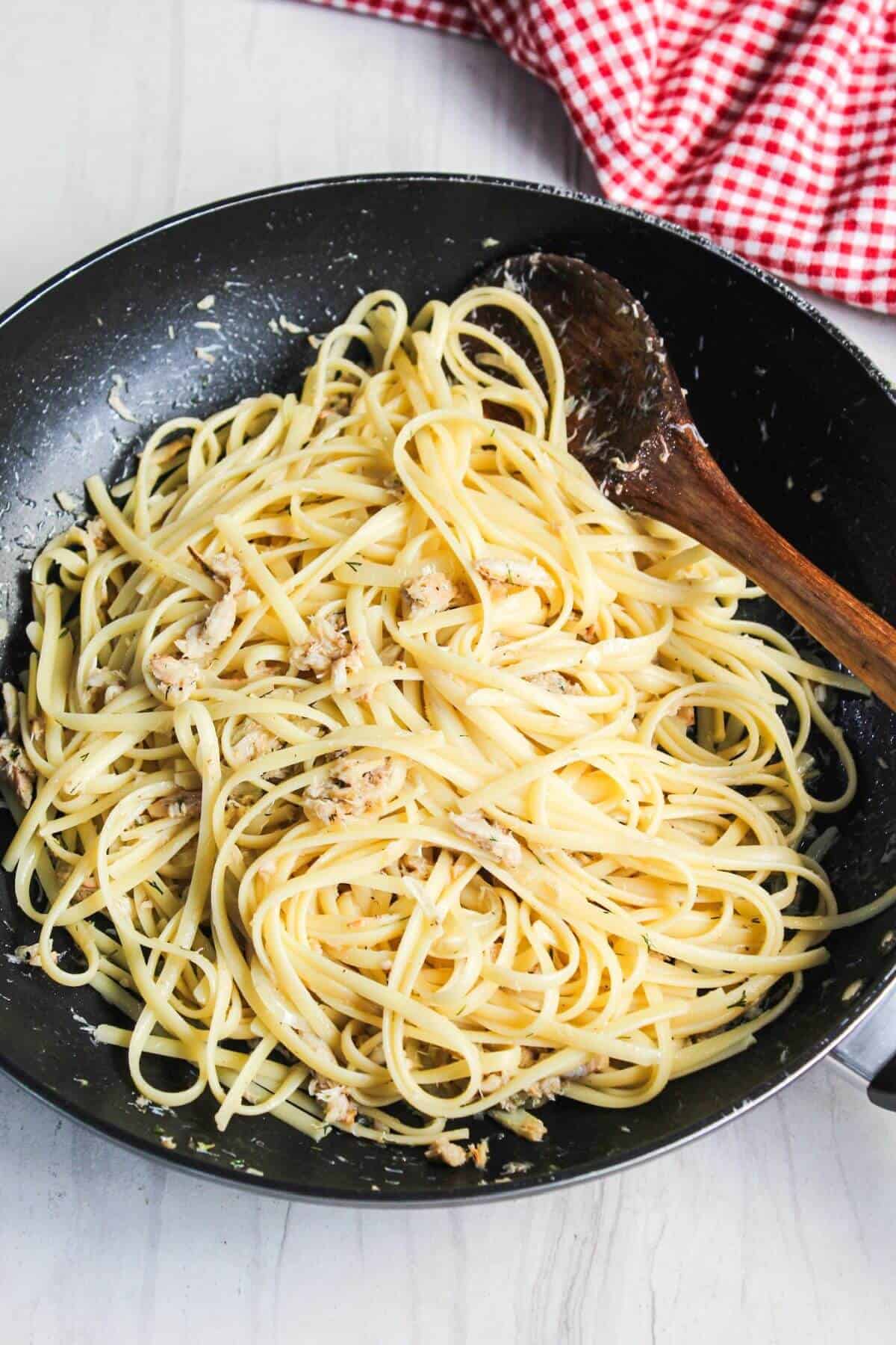 A black skillet containing spaghetti tossed with crab, garlic, and herbs, with a wooden spoon, on a white surface beside a red and white checkered napkin.