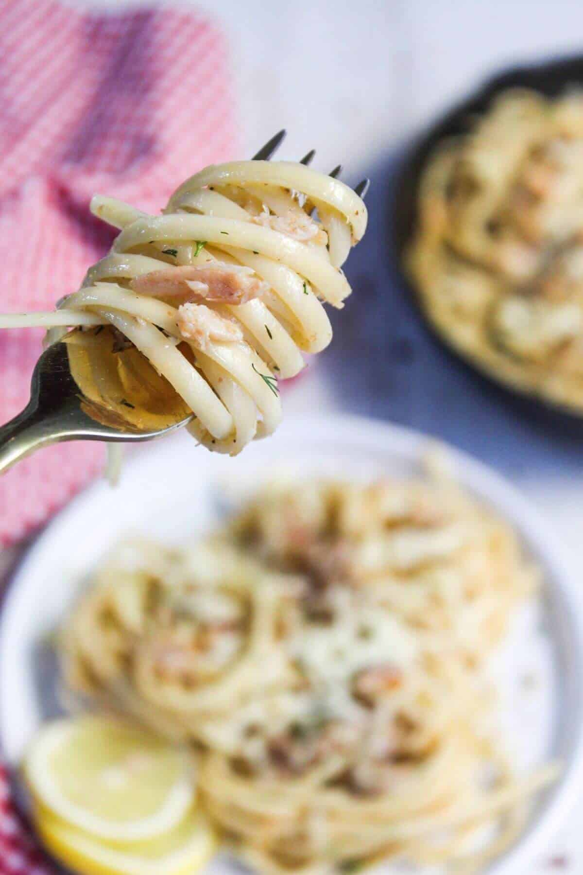 A fork twirling pasta noodles with visible pieces of crab, served on a blurred background with more pasta dishes.