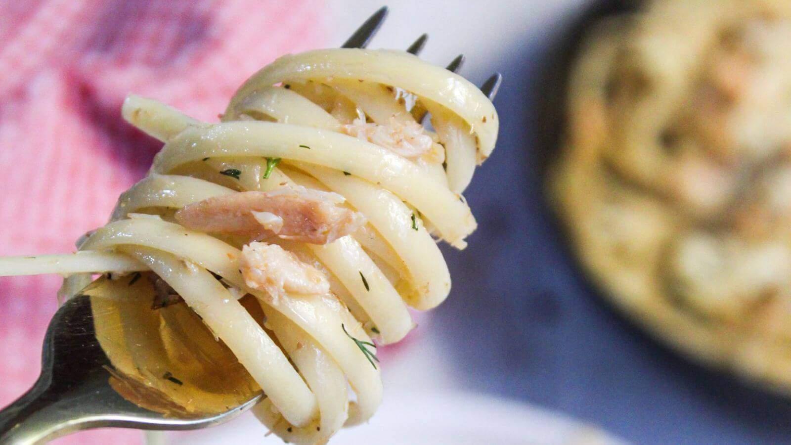 A fork twirling crab pasta with bits of herbs, set against a plate with more pasta and a blurred background.