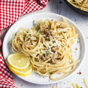 A plate of crab pasta, garnished with parsley and grated cheese, accompanied by lemon slices, on a wooden table with a red checkered napkin.