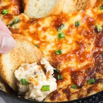 A hand scooping a creamy dip with melted cheese and green onions, served with bread in a cast-iron skillet.