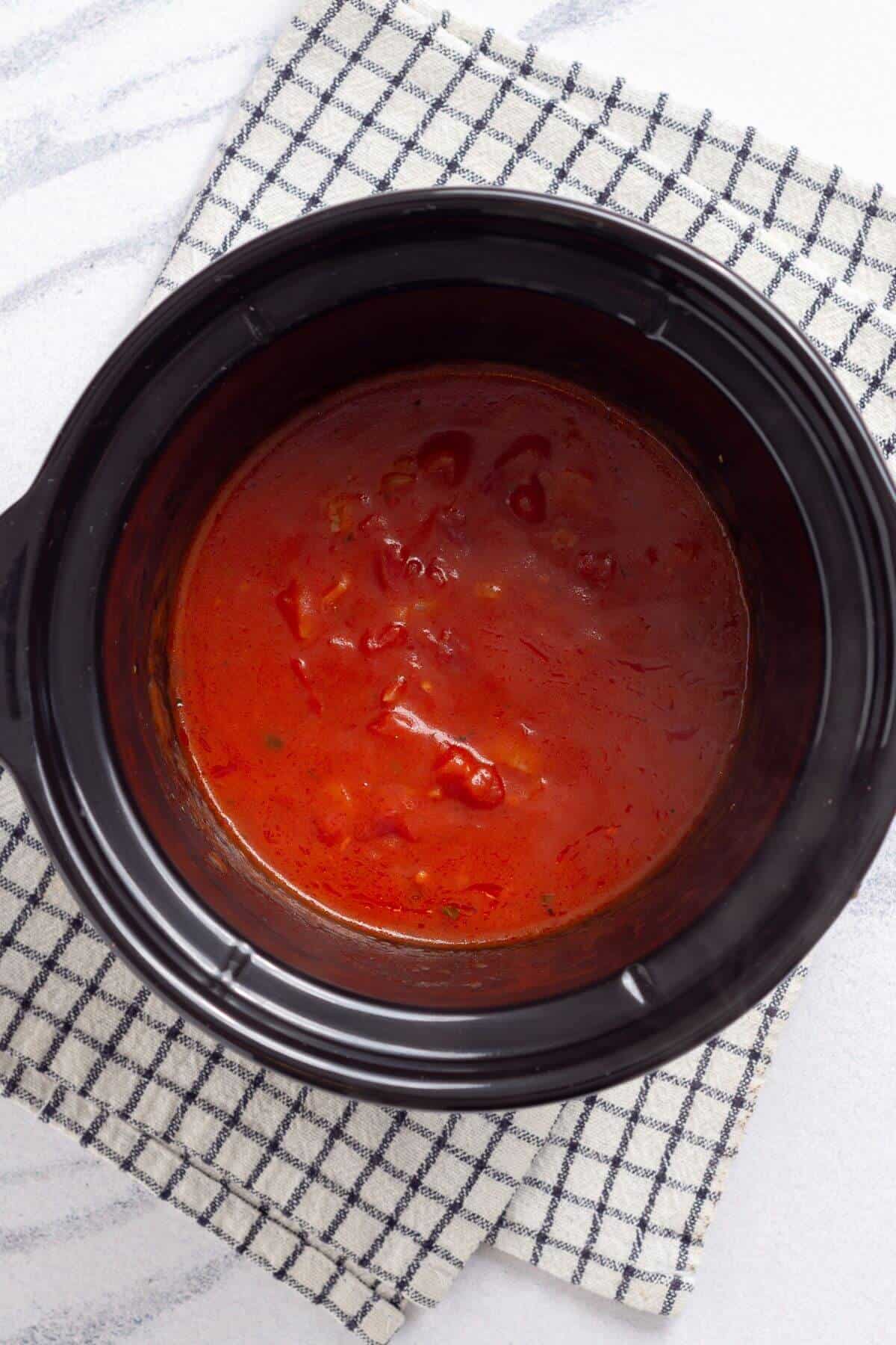 Tomato sauce cooking in a black slow cooker on a checkered cloth.
