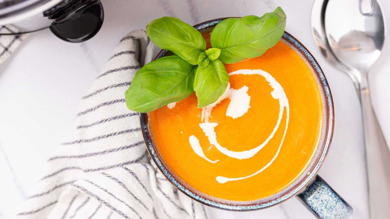 Top view of a bowl of creamy tomato soup garnished with basil, served with a spoon and sunglasses on the side.
