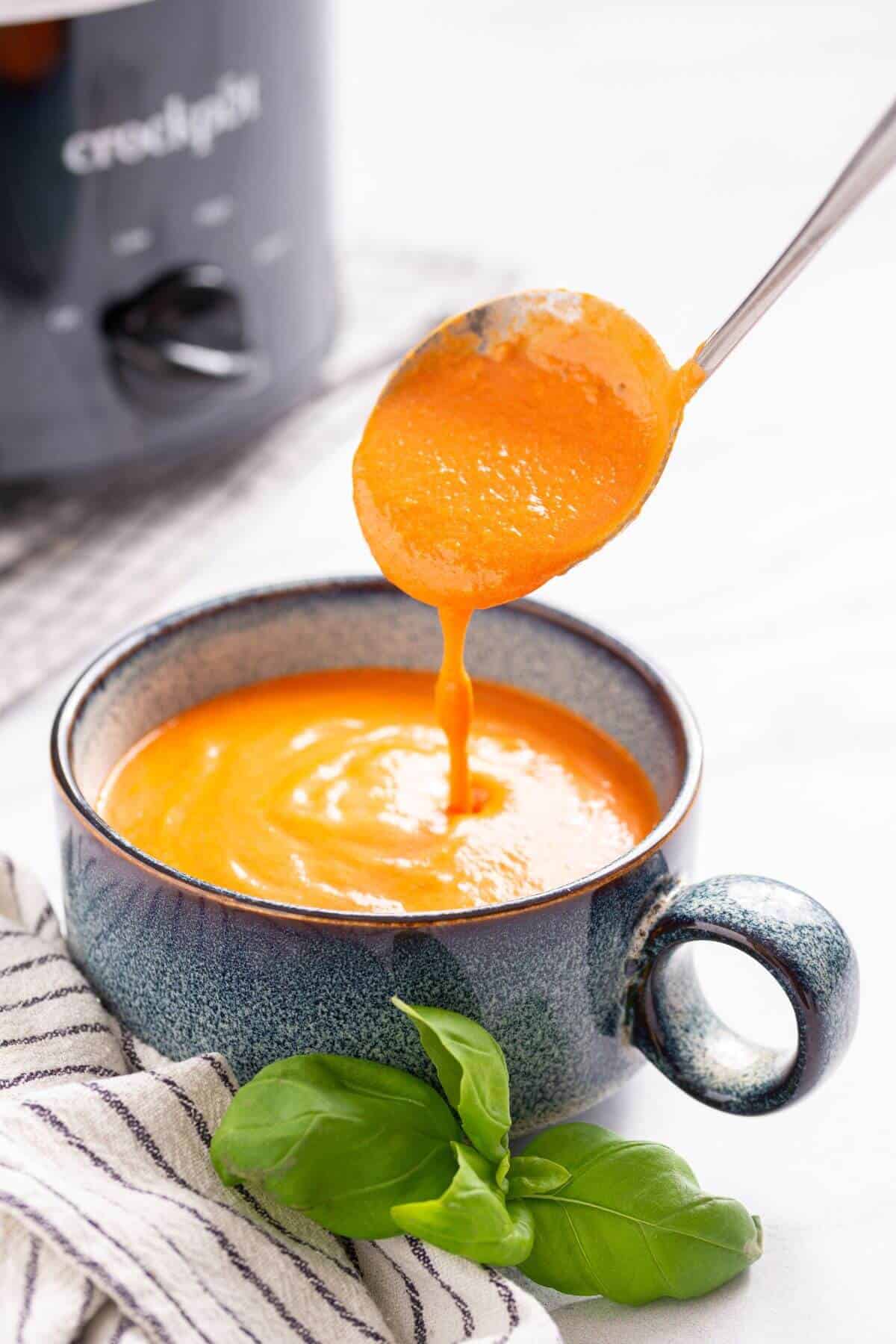 Pouring creamy tomato soup into a blue ceramic bowl, served with fresh basil, near a crock pot.